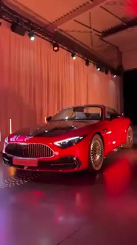 The video shows a red Mercedes-Benz SL Class convertible on display in an event space. The car has a black hood and distinctive silver wheels. A large LED screen displays an image of the same car. The space is decorated with white curtains and illuminated by spotlights. The floor reflects the red and orange lighting. The camera pans from the car to the LED screen and then to the curtains.