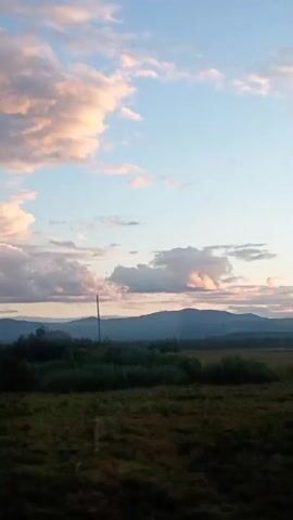 The video shows a wide landscape shot of a grassy field with a distant treeline and mountains under a cloudy sky. The sun is setting, casting a warm glow on the clouds, which are a mix of pink, orange, and blue hues. There is a single utility pole in the middle ground. The camera appears to be moving, possibly from a vehicle, as the foreground shifts slightly.