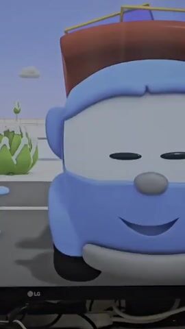 The video is a close-up shot of a blue animated truck character. The truck has large black eyes, a gray oval nose, and a smiling mouth. It is wearing a red hat with a blue brim. In the background, there is a blurry image of a plant and a blue sky with a white cloud. The truck blinks its right eye. The scene then cuts to a wider shot showing the truck from a lower angle, with the same background elements. The truck's body is light blue, and its tires are black. The video also shows a glimpse of some colorful geometric shapes in the background. The bottom of the frame shows the edge of a television screen and some black cables.