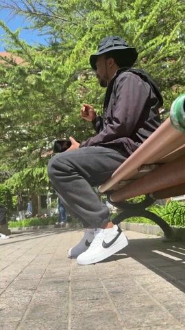 A man sits on a park bench in the daytime. He is wearing a black jacket, gray sweatpants, white shoes with black accents, and a black bucket hat. He holds a phone in his hands. His mouth is pursed, and he gestures with his index finger raised, as if speaking or listening to someone or something. The bench is brown and green. The background features green trees and a building, and the sun is out. The ground around the bench is paved with patterned tiles.