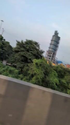 The video shows a perspective from a vehicle moving along a road, with a large industrial structure visible in the background. The structure appears to be a tall, cylindrical silo or processing plant with multiple interconnected sections. It is supported by a metal framework. Surrounding the industrial site are green trees and bushes, suggesting a natural or semi-natural environment adjacent to the industrial area. Several shipping containers, some blue and some yellow, are also visible, indicating a possible logistics or port-related facility. A light pole is present near the structure, and the sky above is cloudy or overcast, with the sun setting or rising, casting a soft light. The foreground of the video is dominated by a gray concrete barrier, part of the road or overpass. The camera is tilted, giving a slightly angled view of the scene.
