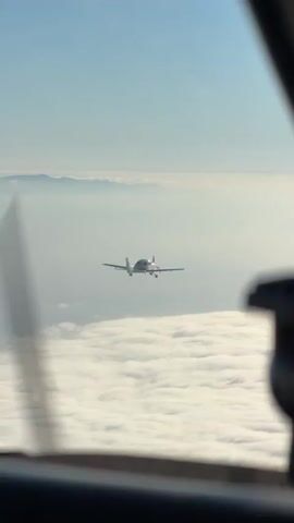 The video shows a view from inside a cockpit, looking out at another small aircraft flying in the distance. The foreground is blurred, with parts of the cockpit visible, including what appears to be a propeller blade that is also blurred from motion. The sky is clear and blue, with a layer of white clouds below the other aircraft. The distant aircraft is silhouetted against the sky and appears to be flying at a similar altitude. There are mountains visible in the far distance, shrouded in haze. The overall impression is that of two aircraft flying in formation or in close proximity to each other above the clouds.