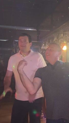 Two men are dancing in a bar. One man wears a white polo shirt and the other wears a navy blue polo shirt with a small pattern. They are both smiling. There are laser lights in the bar. Behind them is a bar counter with glasses hanging above. There is a sign behind the bar and other people are visible further back.