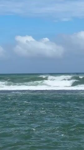 A daytime view of the ocean in Walgama, Sri Lanka, shows large waves breaking. The water is a deep green, and the waves crest with white foam, crashing over a submerged rock or reef formation in the foreground. The horizon is visible, meeting a light blue sky dotted with scattered white clouds. The sound of strong wind and crashing waves is prominent throughout the video.