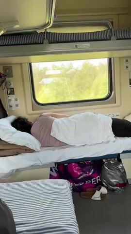The camera slowly pans up and to the right, showing the interior of a train sleeping compartment. The compartment contains two sets of bunk beds on either side of a window. A woman is lying down, facing the window, on the lower bunk on the left, covered with a white blanket. Various bags and a pair of sandals are stored under the bed. Another woman is lying down on the opposite lower bunk, facing away from the window. The upper bunks are folded up.
