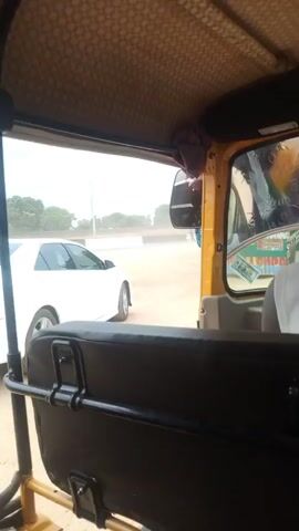 The video begins with a view from inside a yellow auto-rickshaw, looking forward through the windshield. A white car is visible to the left, parked on a dusty, unpaved road. A person's hands are seen in the lower left corner of the frame, gripping a metal bar inside the auto-rickshaw. The scene then shifts to show a person on a motorcycle driving away from the auto-rickshaw. The motorcycle is red and the rider is wearing a light-colored garment. Dust is kicked up by the motorcycle's movement. The auto-rickshaw continues to move, and the white car is again visible to the left, now in motion. The video ends with a view of the white car from the auto-rickshaw. The environment appears to be an outdoor, possibly rural or semi-urban setting, with dry ground and some vegetation in the distance. The sky is overcast.