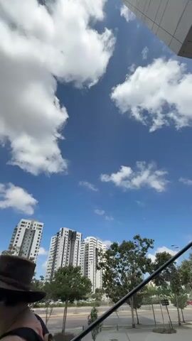 This is a vertical video looking up at a bright blue sky filled with fluffy white clouds. Below the sky, a line of modern, tall residential buildings is visible, interspersed with trees. The architecture of the buildings is contemporary, featuring white and grey facades with many windows. The trees are green and leafy, suggesting a pleasant season. A street with a few cars is visible in the foreground, running horizontally across the lower part of the frame. The scene appears to be an urban or suburban setting in Hod HaSharon, Israel, on a clear, sunny day. The overall mood is peaceful and suggests a typical daytime view. There is no visible text in the video.