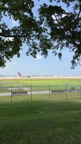A tree with green leaves frames the top of the video. In the middle ground, a chain-link fence separates a grassy area from a flat, open field. Beyond the field is an airport runway with an American Airlines airplane visible. The airplane is white with red and blue accents on its tail. Other airport structures and buildings are visible in the distance. The sky is blue with a few scattered clouds. The video pans slightly to the right throughout its duration.