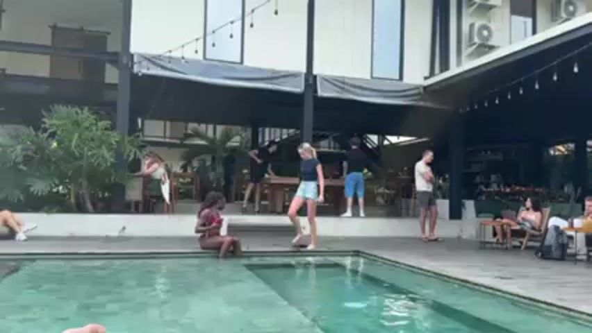 The video shows an outdoor patio area with a swimming pool. Two women stand at the edge of the pool. One wears a bikini and the other a t-shirt and shorts. They appear to be talking to one another. Behind them, people are seated at tables under a covered patio attached to a two-story building. String lights are hung around the patio. Other people are seen walking around or standing near the patio. The swimming pool is green and turquoise.