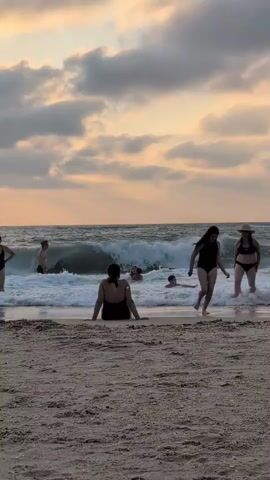 A group of people are enjoying the ocean waves on a sandy beach during what appears to be sunset. The sky is filled with dramatic clouds in shades of orange and gray. Several individuals are in the water, some swimming and others wading. A woman in a black swimsuit is sitting on the sand in the foreground, facing the ocean. To her right, two women in bikinis are walking out of the water. Further out in the ocean, a man in dark shorts is standing, and other figures are visible amidst the waves. The water is choppy, with significant wave action. The sand in the foreground is damp and textured. The overall atmosphere suggests a recreational and relaxed beach scene.