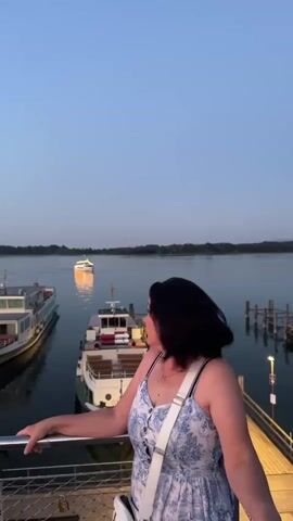 A woman is standing on a dock overlooking a body of water. She is wearing a blue and white patterned dress and a white crossbody bag. She turns to face the camera and smiles. There are several boats docked in the water, and another boat is in the distance with lights on. The sky is a clear, deep blue, suggesting it is either early morning or late evening. The water is calm and reflects the lights of the distant boat. The scene appears to be a peaceful and scenic waterfront location.