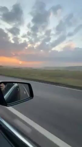 The video depicts a view from a moving car, showcasing a landscape at sunset. The car is traveling on an asphalt road marked with a white line. To the right of the road stretches a green field, with a subtle fog-like layer in the distance where the sun is setting, creating a soft orange glow against the cloud-filled sky. The car's side mirror is visible, reflecting the interior of the vehicle, including what appears to be a hand. As the car moves, trees without leaves pass by on the side of the road, silhouetted against the colorful sky. The overall tone is peaceful and serene.