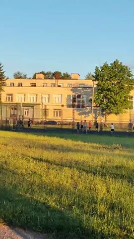 The media file shows a wide shot of a schoolyard in Sumy, Ukraine, during the late afternoon or early evening. The sun casts long shadows from a large tree on the right side of the frame. The sky is a clear, bright blue.

In the background is a pale yellow, two-story school building with a flat roof and many windows. The building appears to have several entrances with covered walkways. A tall chain-link fence separates the building from a grassy field in the foreground.

On the field, behind the fence, a group of children are playing. They appear to be kicking a ball, possibly playing soccer. Some of the children are wearing t-shirts and shorts, while others are in darker clothing. There are at least six children visible, spaced out across the field.

The foreground is dominated by a field of tall, green grass, which is partially lit by the setting sun. The grass is uneven and appears to be a natural meadow rather than a manicured lawn. A dirt path is visible at the very bottom of the frame.

The overall atmosphere is one of relaxed, outdoor recreation. The children are engaged in playful activity, and the warm sunlight suggests a pleasant day. There is no visible text within the media file.