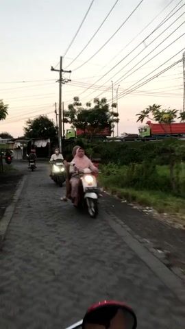 The video shows a street scene at dusk. In the foreground, the red mirror of a motorcycle is visible. Ahead, a person rides a white scooter, with a passenger behind them. Both individuals appear to be wearing light-colored clothing and head coverings. Further down the road, another scooter is seen, also with two occupants. The road is paved with dark stones. On either side of the road, there is overgrown green vegetation and trees. In the background, a utility pole with numerous wires stretches across the frame. Beyond the vegetation, large green trucks are visible on what appears to be a highway. The sky is a muted orange and grey, indicating sunset. The overall impression is of a casual evening ride in a semi-rural or suburban area.