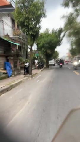 This video captures a street scene in Pejeng, Indonesia, likely during the day as indicated by the lighting. The weather appears overcast with a cloudy sky, suggesting it might be humid or a prelude to rain. The camera perspective is from a moving vehicle, likely a motorcycle or scooter, offering a first-person view of the road ahead.

The scene is characterized by a moderately busy street. The subjects include several people riding motorcycles and scooters, moving in the same direction as the camera. There are also parked motorcycles on the side of the road. The objects visible are various vehicles, including cars, motorcycles, and a truck in the distance. The setting is urban or semi-urban, with buildings lining one side of the street, interspersed with trees and vegetation. The architecture of the buildings appears modest, with some structures having multiple stories.

The primary activity is traffic flow on a street. There are no specific events or gatherings depicted, just the normal movement of vehicles and people in an Indonesian town. Notable details include the presence of lush green trees, which are a common sight in tropical regions like Indonesia. The blurriness of the footage, due to the motion and possibly low resolution, gives the impression of a fast-paced environment. No visible text is discernable in the video. The overall mood is one of everyday life and transit, with a sense of mild congestion.