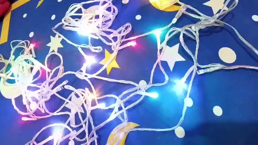 The video shows a tangled string of fairy lights on a dark blue fabric with yellow and white star patterns and white circles. The lights are illuminated in various colors, including white, blue, pink, and green, and they flicker on and off. The camera angle is a top-down view, and the lighting is dim, highlighting the illuminated lights. The scene is static, with no visible people or actions.