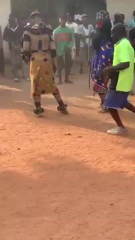 The video shows a group of people participating in a traditional dance or ceremony in an outdoor setting, likely a village or rural area, evident by the red dirt ground. The crowd encircles the central action, which involves dancers in traditional clothing and masks. Some people in the crowd are playing percussion instruments. The dancers are engaged in fast, rhythmic movements, kicking up dust from the ground. The mood appears festive and lively.