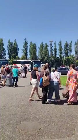 This media file captures a sunny day outdoors, likely in the afternoon given the bright sunlight casting distinct shadows. The scene is an open paved area, possibly a parking lot or a staging area near a transportation hub, with a line of parked buses in the background. Tall, slender trees line the perimeter of the area, suggesting a location with some landscaping or natural surroundings. The sky is a clear, bright blue, indicating fair weather.

The primary subjects are a group of people, appearing to be travelers, engaged in the activity of disembarking from or preparing to board buses. They are diverse in age and gender, and many are seen with luggage, including suitcases on wheels, indicating travel. The individuals are walking, some alone and others in small groups, suggesting a movement or transition.

There are no discernible emotions visible due to the distance and quality of the image. The activity appears to be orderly, with people moving through the space with their belongings. The presence of multiple buses and the luggage strongly suggests an organized trip, perhaps a tour group or a charter service.

The objects in the scene include several buses of varying colors, cars parked in the background, lampposts, and various pieces of luggage. A circular manhole cover is visible in the foreground. No visible text is present on the buses or in the immediate surroundings that would provide specific location or destination information. The overall impression is one of a busy but uneventful moment of travel logistics.