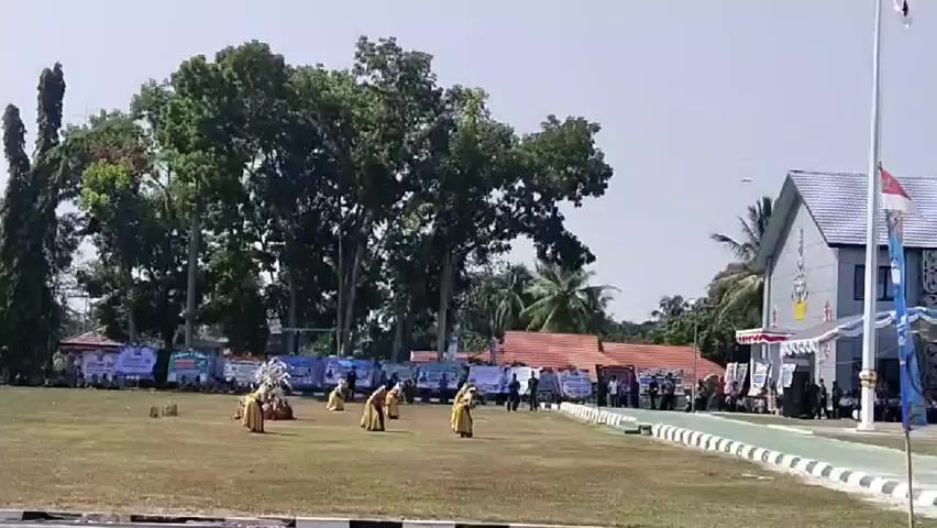 A wide shot shows an outdoor event on a grassy field. In the foreground, several people in yellow and gold traditional attire are performing a dance. In the background, there are rows of people seated and standing, with banners and tents set up. Behind the seated spectators, there are several trees and buildings. The sky is clear and bright. The overall impression is of a community gathering or performance.