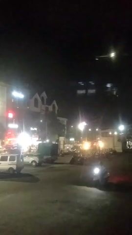 The video shows a night scene of a city street. Several cars and motorcycles are parked or moving, and there are buildings with lights on.  There's a traffic light and streetlights illuminating the scene. People are visible in the distance. The overall impression is of a typical urban night setting.