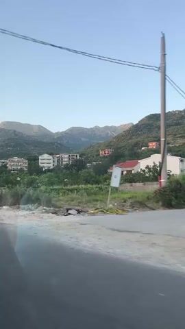 This video captures a view from a moving vehicle, likely a car, driving along a road in Podlastva (Poljice), Montenegro. The scene unfolds during what appears to be the late afternoon or early evening, given the soft, golden light bathing the landscape. The sky is clear and a pale blue, suggesting fair weather.

The foreground is dominated by the road and a gravelly shoulder. Beyond this, the landscape rises into rolling hills and mountains covered in lush green vegetation. Scattered among the greenery are several buildings, including multi-story apartment complexes and individual houses, some with red-tiled roofs. There's a sense of ongoing construction or development in some areas, with unfinished structures visible.

Several cars are parked along the roadside. One notable object is a large, arched gateway constructed from stone. A billboard with text in what appears to be a Slavic language is visible on the left side of the frame, partially obscured by trees. The text includes "lovćen" and "Tehnički pregled."

In the latter part of the video, a person's arm is visible resting on the open window of the vehicle the footage is being filmed from, suggesting a passenger is enjoying the drive and the scenery. The overall mood is peaceful and scenic, showcasing a somewhat rural and developing area with a mountainous backdrop.