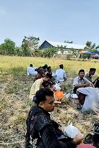 A group of men are sitting on the ground in an outdoor setting, possibly a field or park. They are dressed in traditional Islamic attire, including peci caps. Some are engaged in conversation, while others appear to be eating or resting. There are plastic bags and food items scattered around them. In the background, there is a building, trees, and a clear sky. The overall atmosphere seems relaxed and communal.
