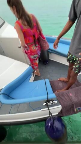A woman with long brown hair walks into a white boat, wearing a brightly colored top and skirt, and carrying a pink mesh bag. The boat has blue and white cushioned seats and black hexagonal patterned floor mats. A man stands on the boat, wearing black shorts and sandals. The person recording the video is also on the boat, wearing black shorts and sandals. The boat is surrounded by green water. The man on the boat says something about “tomorrow.”