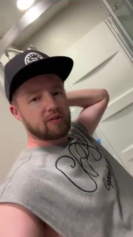 A man wearing a gray t-shirt and a black baseball cap is in a room. He has his right arm behind his head and appears to be looking at the camera. The room has white walls with a textured pattern.