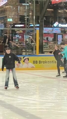 The video shows an indoor ice rink with multiple people ice skating. At the start, a young person wearing a black hoodie with a white star and wide-legged blue jeans is standing in the center. A few other skaters are moving around in the background. Later, a young woman in black is holding hands with a person wearing a blue jacket and dark pants, skating together. Other skaters are scattered around the rink. The rink is surrounded by glass barriers with advertisements and store signs visible behind. The ice is bright and reflective.