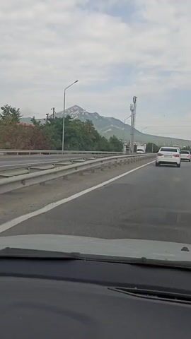 A car is driving down a highway. The road has multiple lanes and a metal guardrail on the left side. Beyond the guardrail, there are trees and a mountain in the distance. A cell tower is visible near the mountain. Several cars and a white truck are also on the highway, driving in the same direction as the car capturing the footage. The sky is cloudy.
