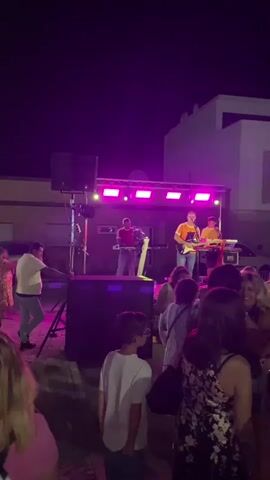 A nighttime outdoor gathering is taking place with a live band performing on a stage. The band consists of at least three musicians, one playing an accordion, one playing a guitar, and another near a keyboard. The stage is illuminated by pink and purple lights. A crowd of people are gathered in front of the stage, some dancing and interacting. Decorations, including colorful banners and a lantern, are visible. The environment appears to be a street or public square with buildings in the background. Several portable toilets are also present. The video shows people of various ages enjoying the event.