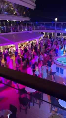 This video captures a lively outdoor evening scene on what appears to be a cruise ship deck. A large crowd of people, dressed casually in summer attire, are gathered and dancing under vibrant pink and purple lights. The atmosphere is festive, with music seemingly playing, judging by the dancing.

In the background, several levels of the ship are visible, including railings and balconies where more people can be seen. One prominent feature is a circular hot tub, illuminated with blue light, with several individuals socializing around it. There's also a bar area with patrons being served, indicated by the presence of a counter and people standing at it. Tables and chairs are scattered across the deck, suggesting areas for relaxation and dining. The night sky is visible above, adding to the nighttime ambiance. The overall impression is one of a party or a nightclub scene taking place on a moving vessel.