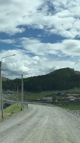 The video shows a gravel road curving to the right, leading towards a village nestled at the base of a lush green, tree-covered mountain. The sky is partly cloudy with patches of blue. Several streetlights are visible on the left side of the road, along with a guardrail. In the distance, houses with dark roofs can be seen scattered throughout the village, and a few animals, possibly cows, are grazing in a field on the right.