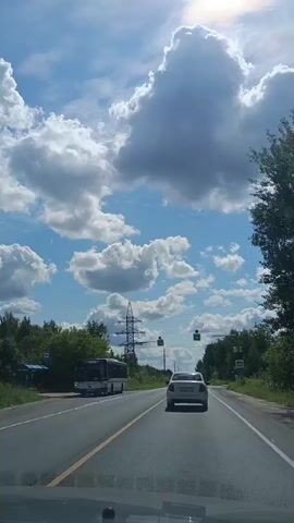 The video shows a car driving on a paved road under a blue sky with scattered white clouds. The sun is visible through the clouds, creating a bright effect. On the left side of the road, there is a bus and some trees. There are also power lines and poles along the road. Traffic lights and pedestrian crossing signs are visible overhead. The road has yellow and white lane markings. The perspective is from inside another vehicle, looking forward through the windshield. The car in front is white.