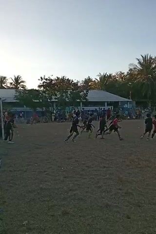 The video shows a group of young people playing soccer on a dry, grassy field. There are spectators in the background, as well as trees and buildings. The players are wearing different colored jerseys, and some are kicking a ball while others are running. The sky is clear and the sun appears to be setting, casting long shadows across the field.