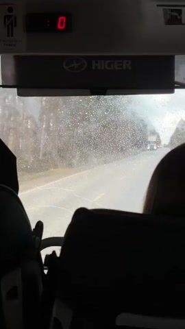 The video captures a first-person view from inside a moving vehicle, likely a bus, looking out through the front windshield. In the foreground, the dark silhouettes of two individuals, presumably passengers, are visible. Above the windshield, a digital display shows numbers changing from "0" to "6", then "1", and back to "0" throughout the video. Below this display, a rectangular device features the "HIGER" logo and text. To the left, a sign displays a "no standing person" icon along with the Russian text "ПРИСТЕГНИТЕ РЕМНИ!" (Fasten your seatbelts!).

The windshield is speckled with raindrops, indicating rainy or recently rainy weather, and the road appears wet. The vehicle is traveling on a road, possibly a highway, with a line of trees visible in the distance along the roadside. Other vehicles, including several trucks and cars, are seen on the road ahead, some approaching and some receding. The scene depicts a journey during daytime under overcast conditions in the Iiul'skoe region of Russia.