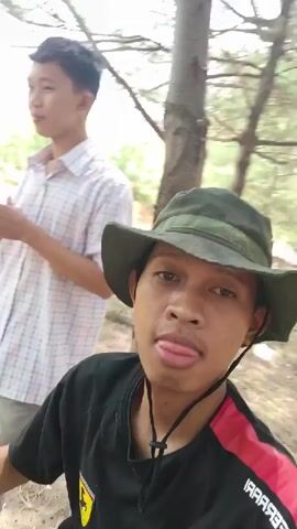 Two young males are outside in what appears to be a wooded area. The male in the foreground is wearing a black t-shirt with a red and white stripe on the right sleeve, and a dark green bucket hat. He is holding the brim of his hat, and then touches the top of it. He opens his mouth and sticks his tongue out slightly. The second male is behind and to the left of the first, wearing a white and light blue plaid shirt and a dark colored strap across his chest. The background shows trees and foliage, with dappled sunlight. The camera is held by the foreground male, giving a selfie-style perspective.