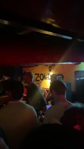 From a ground-level perspective, the video captures a live music performance in a dark, crowded bar or club. The band, consisting of around five members, is visible on a slightly raised platform behind the crowd. The backdrop features a yellow sign with "ZOLDER" written in bold letters. The stage lighting is dim, with some brighter spotlights illuminating the band. The audience is a mix of people, mostly facing the stage. Some are holding up phones to record the performance. The ceiling above is low and dark, with exposed beams and red paint. The atmosphere is lively, with a sense of casual enjoyment.