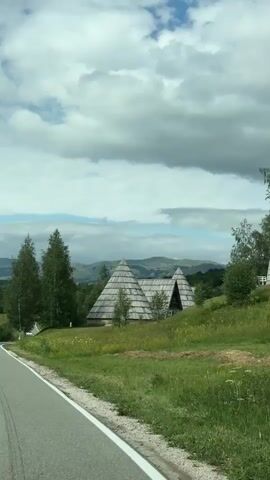 The video shows a car driving down a paved road through a lush green countryside. The road curves to the left, and on the right side of the road, there are two distinctive pyramid-shaped buildings with wooden roofs. Tall, slender trees are visible behind the buildings. Further in the distance, rolling hills and mountains can be seen under a cloudy sky. Several cars are driving on the road, with some passing by the camera's viewpoint and others moving away from it. The overall scene depicts a scenic drive through a rural, possibly mountainous, area.