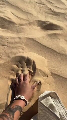 The video shows a person’s hand sifting sand in a desert environment. The sand is light brown and forms small dunes. The person has a bracelet and a tattoo on their arm. They gently lift the sand and let it fall back onto the dune. A piece of light grey fabric is visible next to the hand. The environment appears to be sunny and bright.