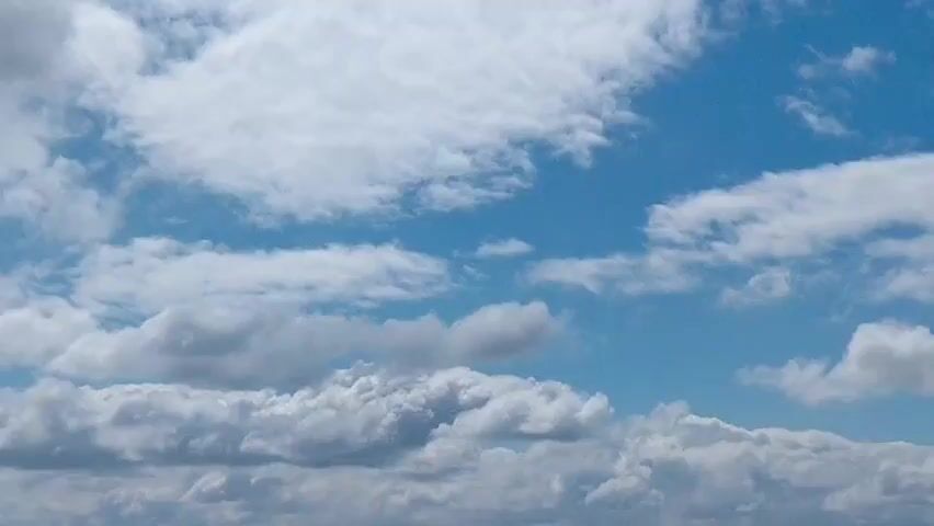 The video shows a wide shot of a bright blue sky with white, fluffy clouds. The clouds are varied in size and density, with some appearing as thin wisps and others as large, billowing masses. The camera pans slightly to the left, revealing a distant landscape of green fields and trees, with a few scattered buildings. The lighting suggests it is daytime, possibly late morning or early afternoon. There are no people or discernible actions captured in the video.