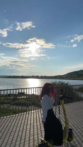 A woman with red curly hair, wearing a white striped shirt and a black skirt, stands on a yellow electric scooter on a paved walkway. She is looking at the camera and smiling. Behind her, a body of water is visible, bordered by a metal fence and trees. The sun is shining brightly in a blue sky with scattered clouds. The scene appears to be a scenic outdoor location, possibly a park or a waterfront area. The video pans to the right, showing more of the walkway and the landscape.
