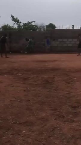 The video shows three young men playing soccer on a dirt field in front of a cinder block wall. One of the men is wearing a green shirt, and the other two are wearing a blue shirt and a black shirt, respectively. The men are barefoot and are kicking the soccer ball back and forth. The environment appears to be an unkempt outdoor space with sparse vegetation and debris scattered on the ground.