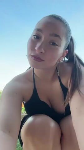 A first-person perspective video shows a young woman crouching outdoors under a clear sky. She is wearing a black bikini top and has her hair pulled back in a ponytail. She makes various facial expressions, including posing with a playful look, smiling, puckering her lips, and looking directly into the camera with wide eyes. There is some foliage and a fence visible in the background.