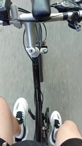 The video begins with a close-up point-of-view shot of a person on a bicycle, showing their legs, feet in white and green athletic shoes, the handlebars, a silver stem, a black bicycle frame, a black chain, and a gear shifter. Attached to the handlebars are a black bicycle light and a silver electronic device, possibly a GPS or speedometer. The camera then pans up and slightly backward to show two people riding bicycles ahead on a paved road. The person in front is wearing a pink shirt and black shorts, while the person behind them is wearing a white t-shirt and dark shorts. The road is bordered by trees and vegetation on both sides. The sun appears to be low in the sky, casting a warm light. The video continues with the point-of-view perspective of the cyclist, showing the road ahead and the two cyclists in front, with the scenery passing by. The road is cracked and weathered, and there is a white line marking the edge of the lane. The trees and bushes are dense and green. The video captures the experience of cycling on a relatively quiet, rural road in what appears to be the late afternoon or early evening.