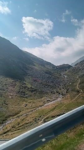 A video shows a mountainous landscape with a winding road. A car is visible on the road. There are buildings along the road and some trees. The sky is blue with some clouds. The overall impression is a scenic drive through a mountain pass.