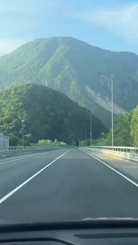 A POV shot from a car driving on a highway with a mountain range in the background. The highway has two lanes in each direction separated by a white line. There are streetlights on the right side of the road and a metal guardrail. The mountains are green and covered in trees. The sky is blue and clear. A small white structure, possibly a tower, is visible on the side of one of the mountains.