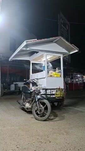 A man in a patterned shirt and tan pants approaches a food cart that appears to be attached to a motorcycle. The cart is white with a peaked roof and appears to be selling food, possibly corn, given the yellow items visible inside. The man leans over the cart, interacting with something inside, and the scene is illuminated by artificial lighting in what appears to be an outdoor setting at night. The motorcycle is parked in front of the cart.