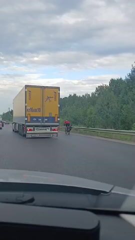 The video shows a road scene from the perspective of a car. A large yellow semi-trailer truck with "KZ16ABE10" and "SCHMITZ CARGOLL" written on it is in the foreground, driving on the right side of the road. Behind the truck, a cyclist in a red shirt and black shorts is riding a bicycle, also on the right side of the road. The road appears to be a highway with multiple lanes, bordered by a guardrail and trees on the right. The sky is overcast with clouds, suggesting it could be a cloudy day. Other vehicles are visible in the distance on the road. The time of day appears to be daytime, possibly late afternoon or early evening, given the lighting. The overall scene depicts traffic on a highway in what appears to be a rural or suburban setting.