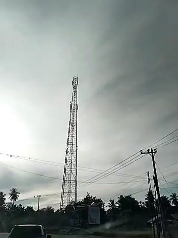 The video captures a view from a vehicle driving on a road. In the distance, a tall, lattice telecommunications tower stands against a cloudy sky. Electrical wires stretch across the frame, supported by utility poles. The landscape features palm trees and other vegetation. Sunlight breaks through the clouds, creating a bright glare. Two cars are visible on the road, one in the foreground and another further ahead. The camera perspective suggests the video is being recorded by someone inside the moving vehicle.